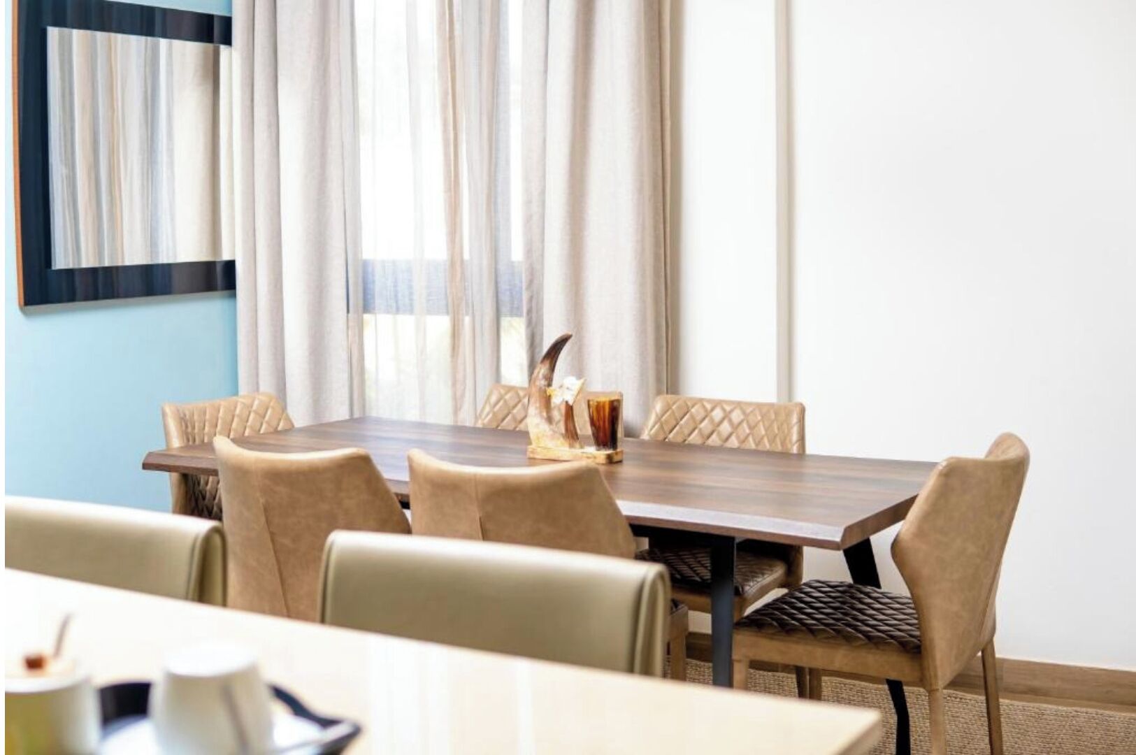 Een hotel eetruimte met een rechthoekige tafel, zes gestoffeerde stoelen en een raam met transparante gordijnen.