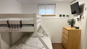 Una litera con escritorio y cómoda, una ventana con cortinas, y un televisor en la pared.