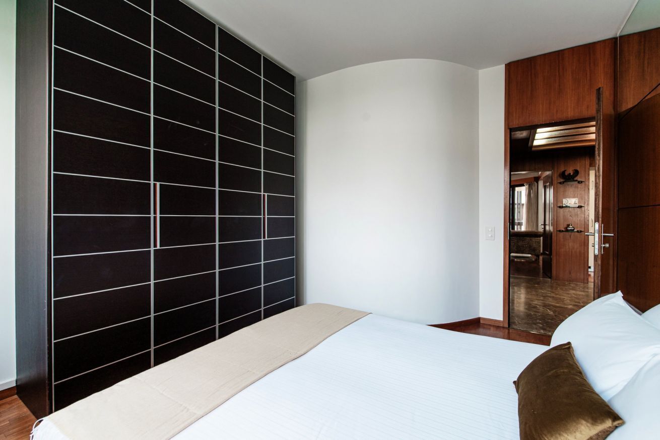 A bedroom with a bed, a black tiled partition, a wooden wardrobe, and a doorway leading to a bathroom.