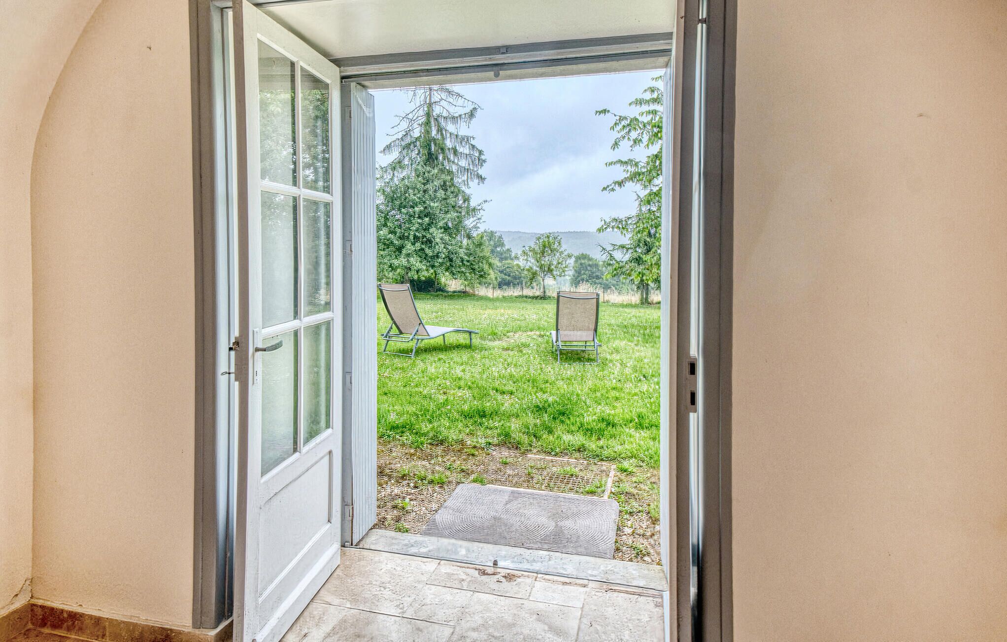 Een open deur die leidt naar een grasveld met twee stoelen en een gestructureerde mat bij de ingang.