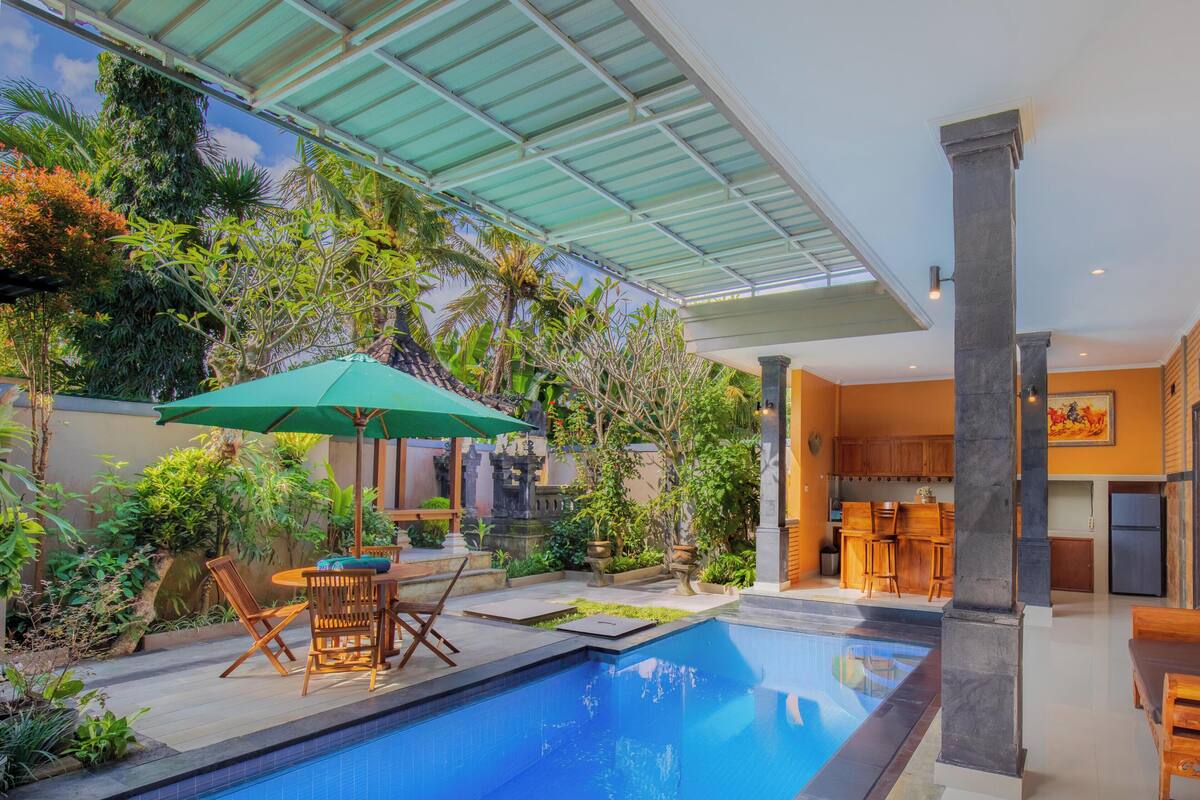 A swimming pool with a green umbrella, wooden furniture, and a bench with cushions.