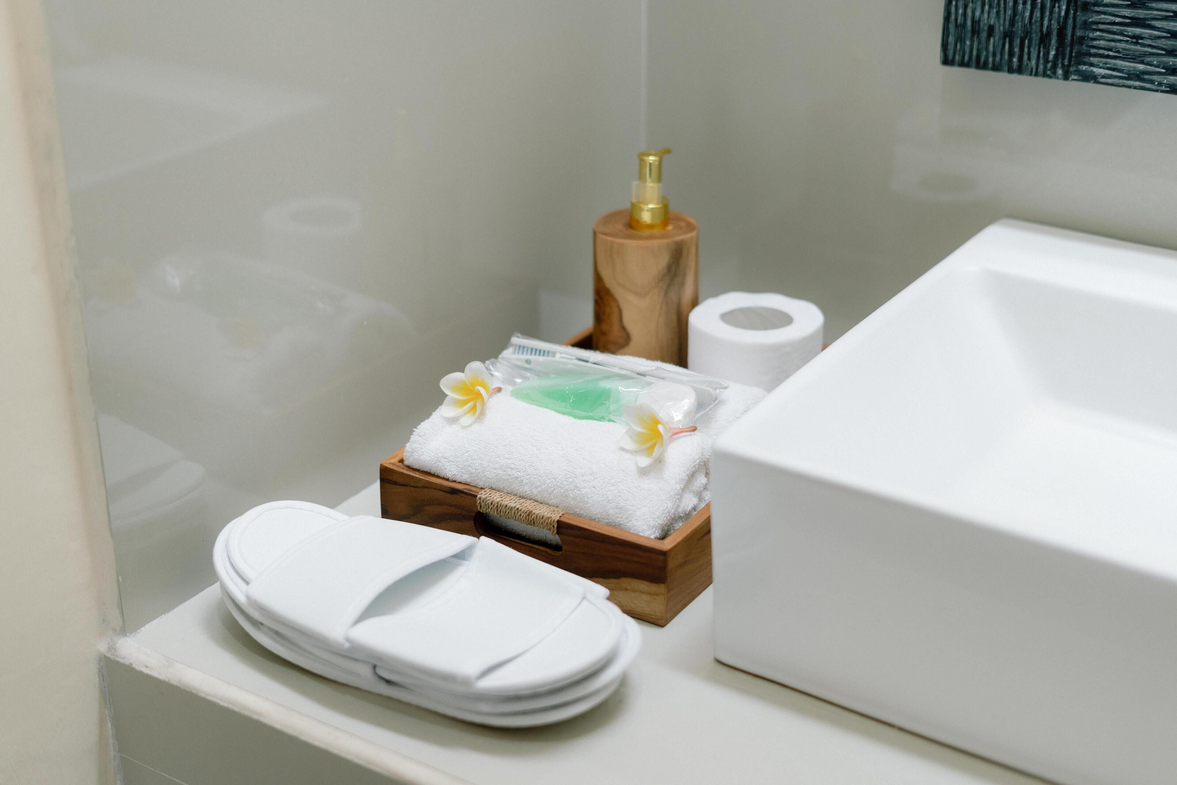 A bathroom with a white sink, a wooden soap dispenser, a roll of toilet paper, and a small wooden box containing a towel and a bottle of lotion.