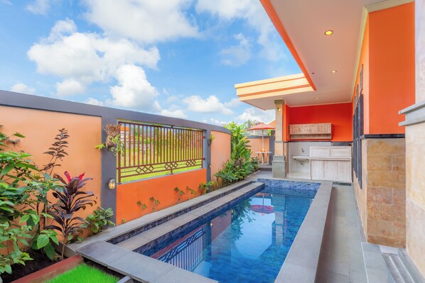 A swimming pool with a stone-lined edge, surrounded by a garden with various plants and a metal gate. - Moning Villa Ubud (Ubud)