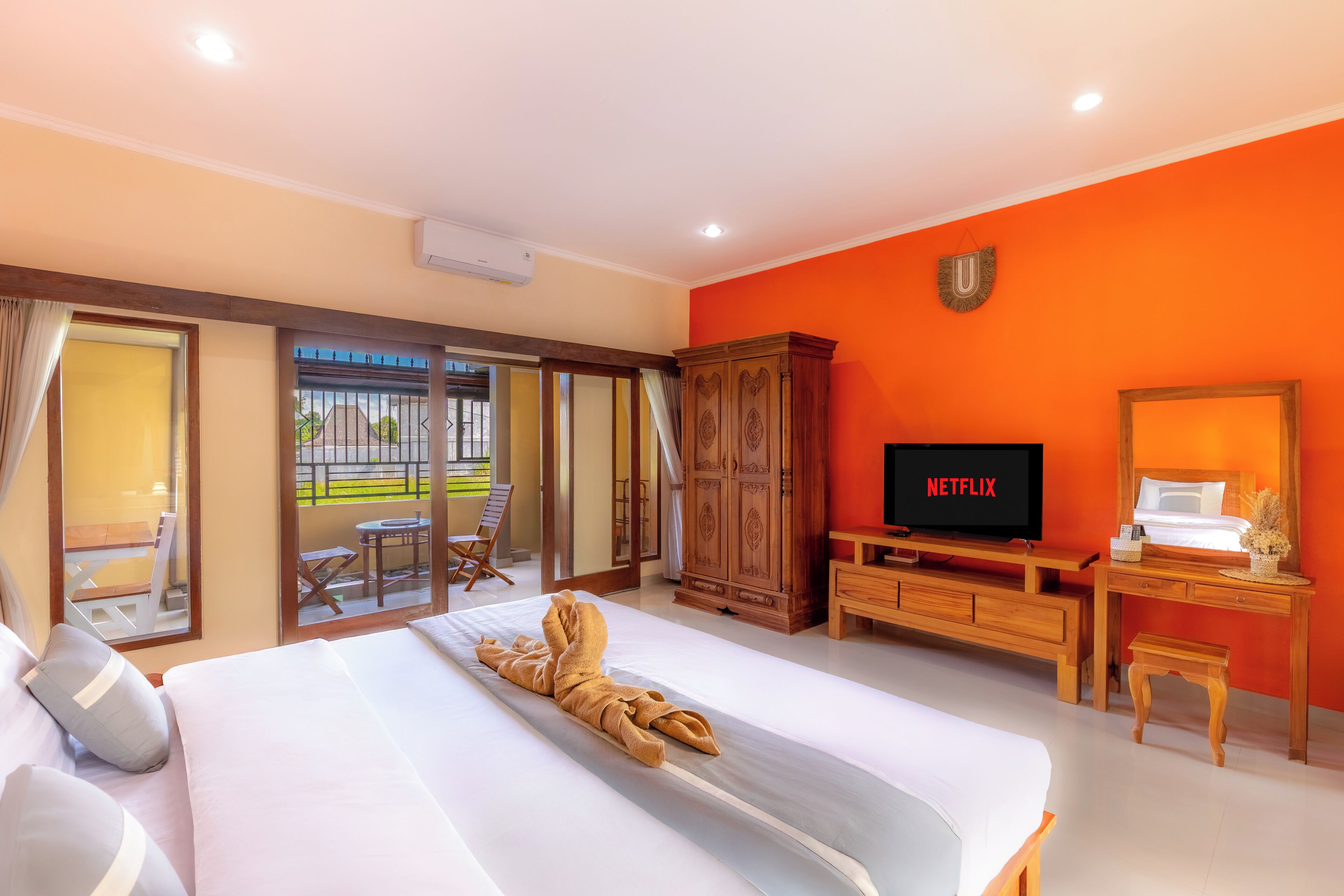 A hotel room with a large bed, a wooden TV stand with a Netflix logo, a wooden cabinet, and a balcony with outdoor seating.