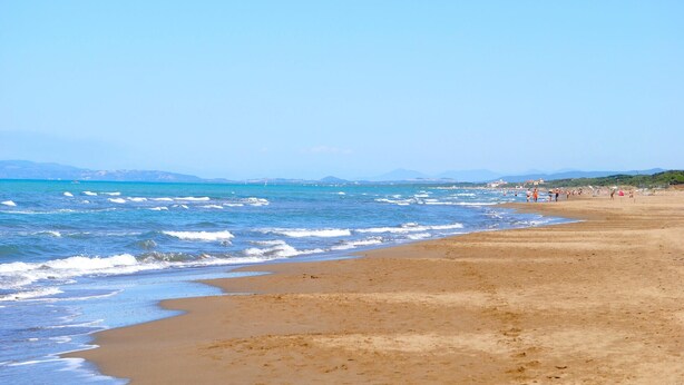 A wide sandy beach with gentle waves, a clear sky, and distant mountains.