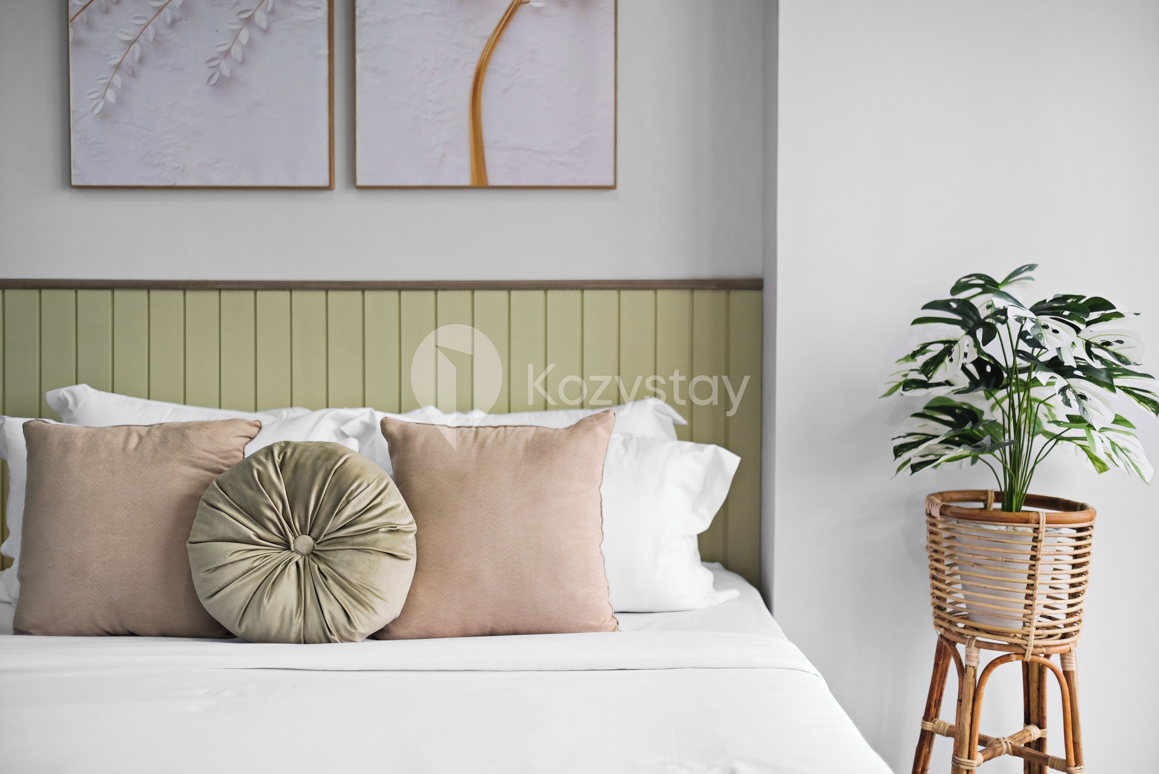 Una cama bien hecha con almohadas blancas y beige, un cabecero verde y un atril de mimbre con una planta verde.