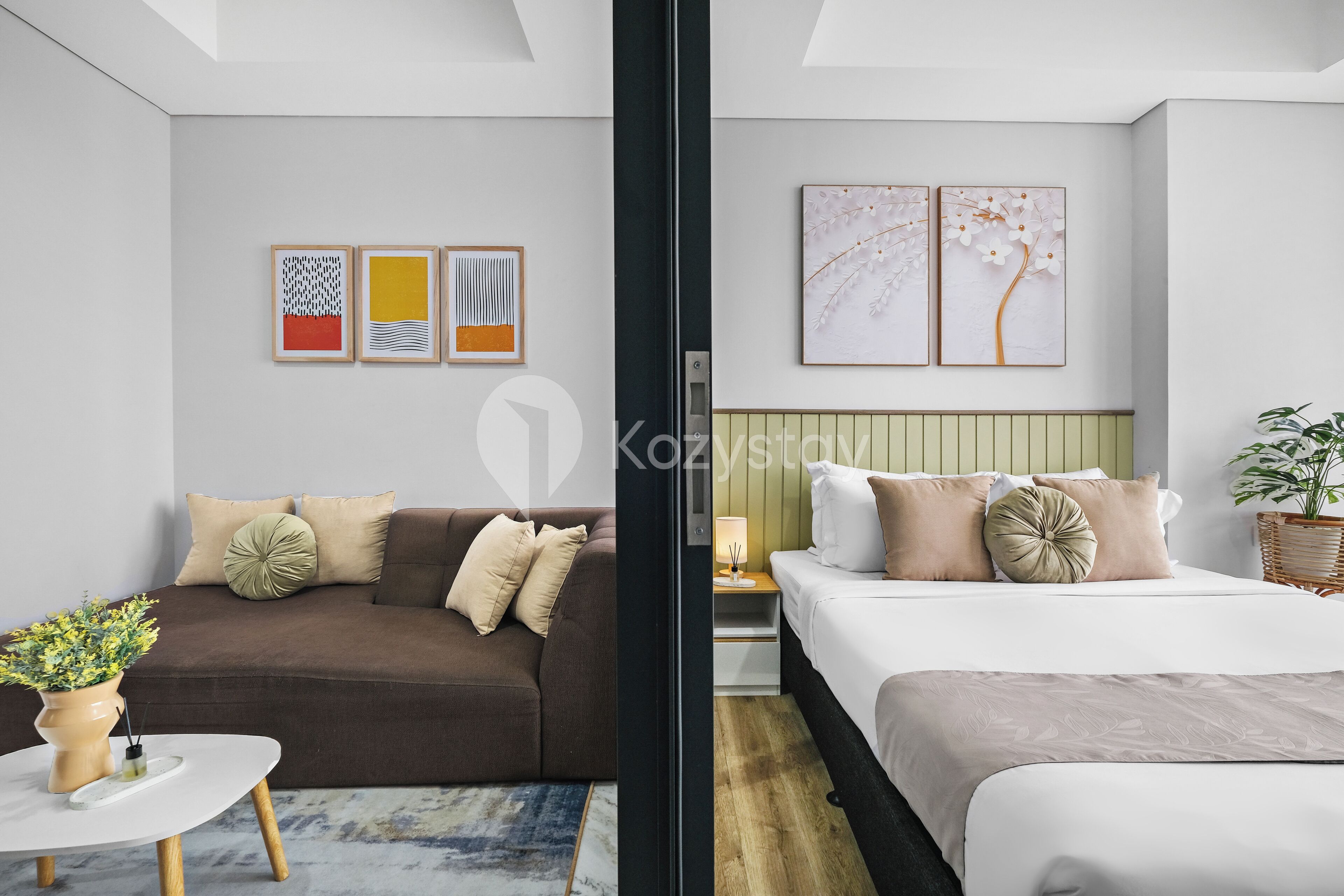 Quarto de hotel moderno com sofá castanho, cama branca e uma pequena mesa com uma planta.
