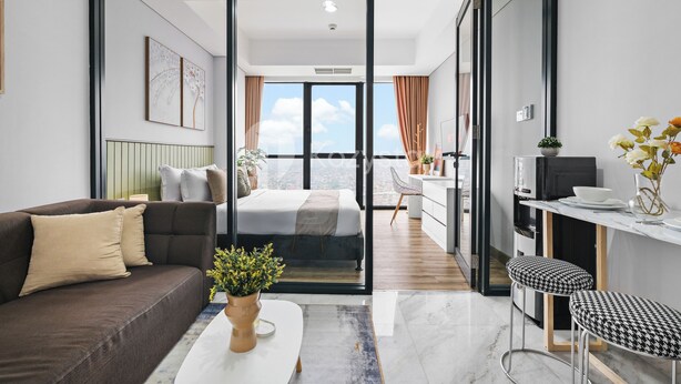 A modern apartment with a large sliding glass door leading to a balcony, a sofa, a bed, a kitchen area, and a dining table with bar stools. - Branca by Kozystay - Alam Sutera (Tangerang)