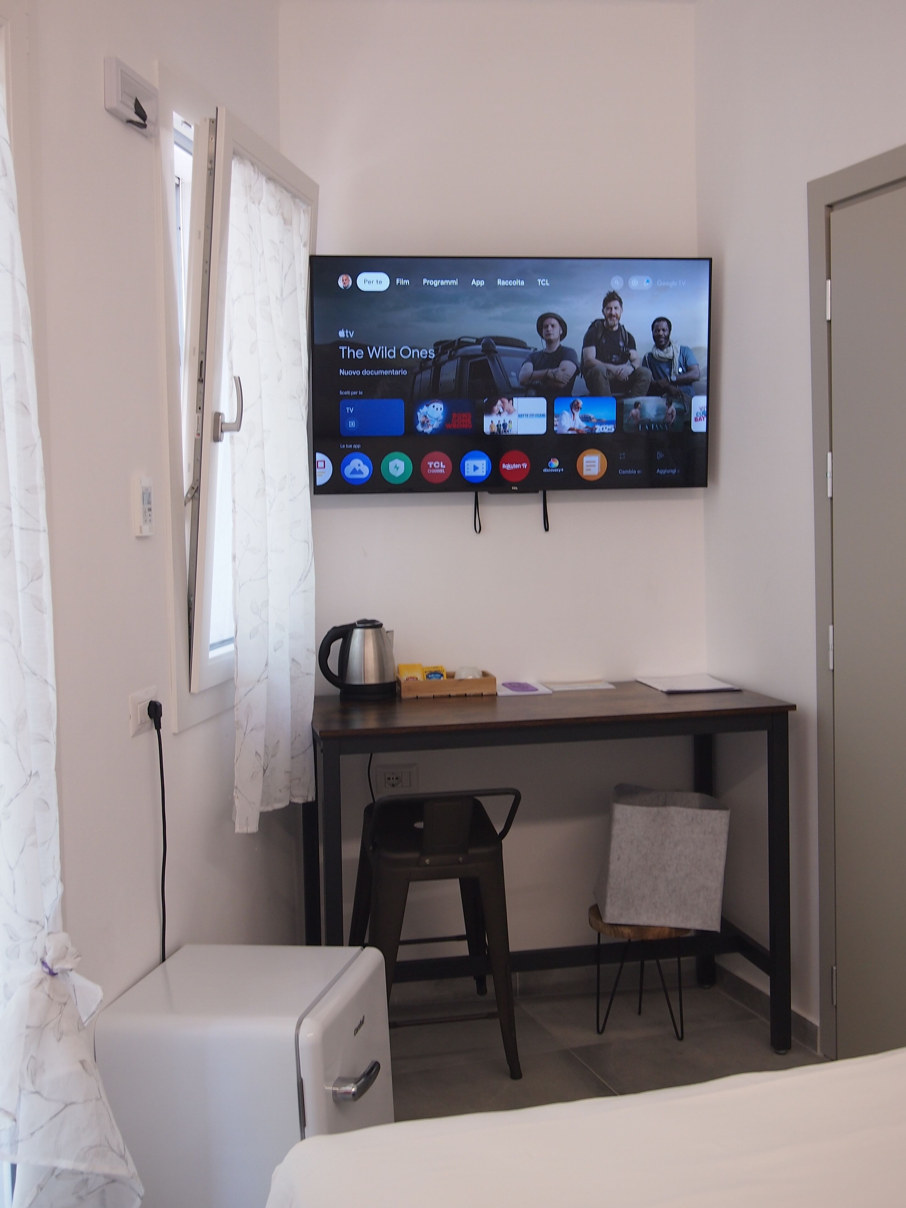 A hotel room with a flat-screen TV mounted on the wall displaying a documentary. There is a desk with a kettle, a chair, and a small table with a tray of items. A white refrigerator is visible in the corner.