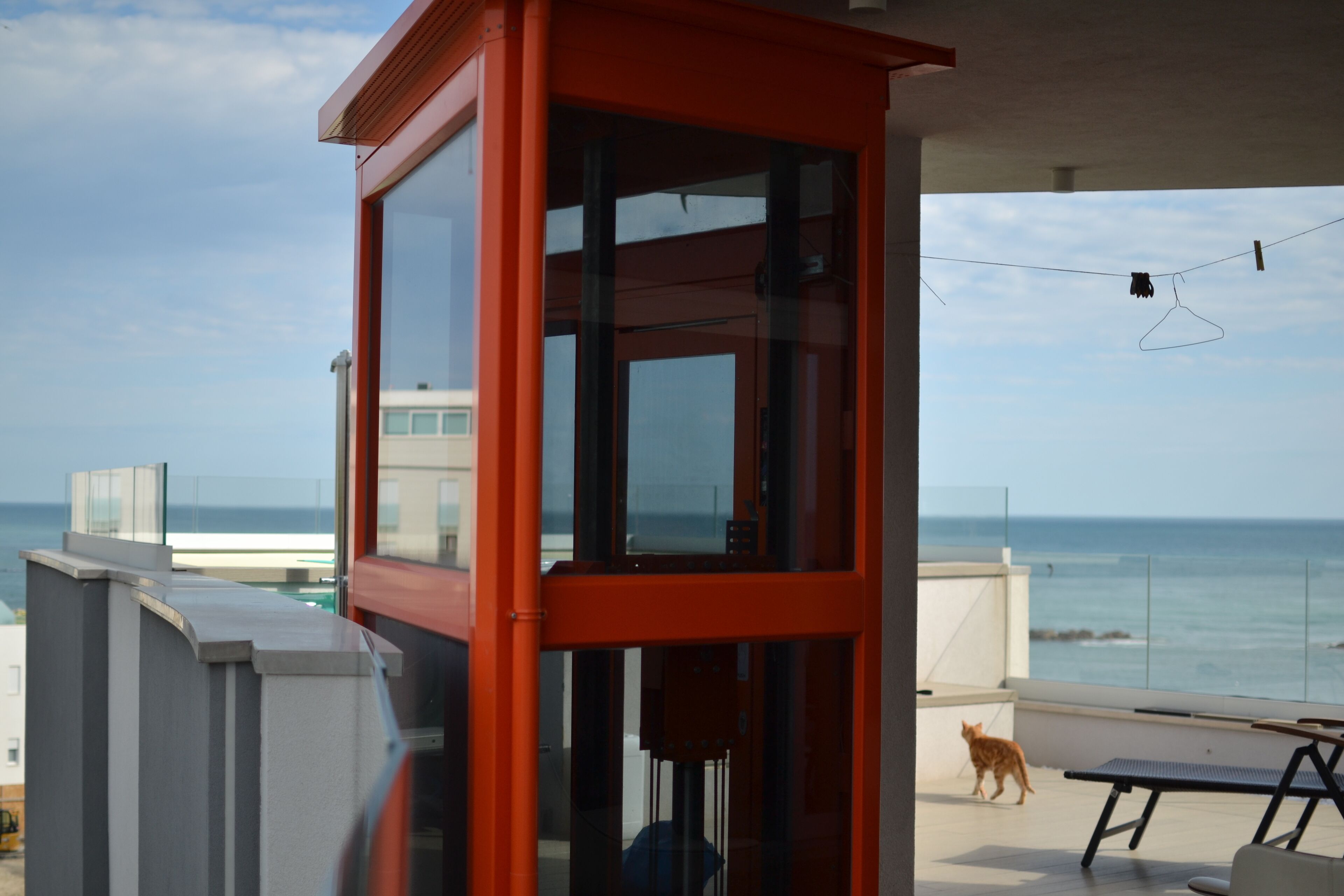 A balcony with a glass enclosure, a view of the ocean, and a cat walking.