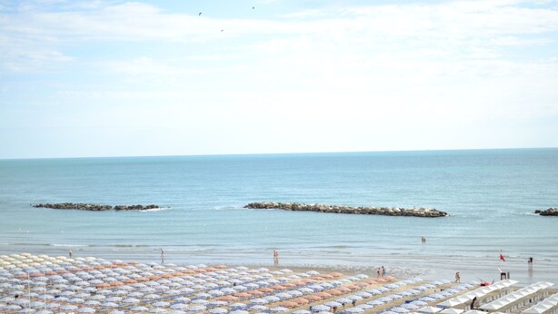 Una playa con numerosos sombrillas y sillas de descanso, un rompeolas rocoso y mar tranquilo.
