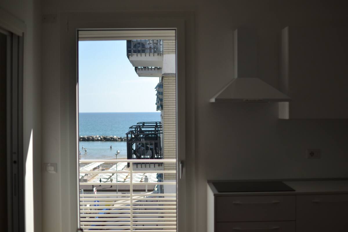 Vista de um quarto com uma janela grande que dá para uma praia com barcos e céu claro.