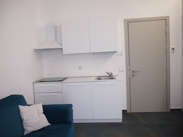 A compact kitchenette with a sink, cabinets, and a stove. There is a teal sofa with a patterned pillow and a closed door in the background.