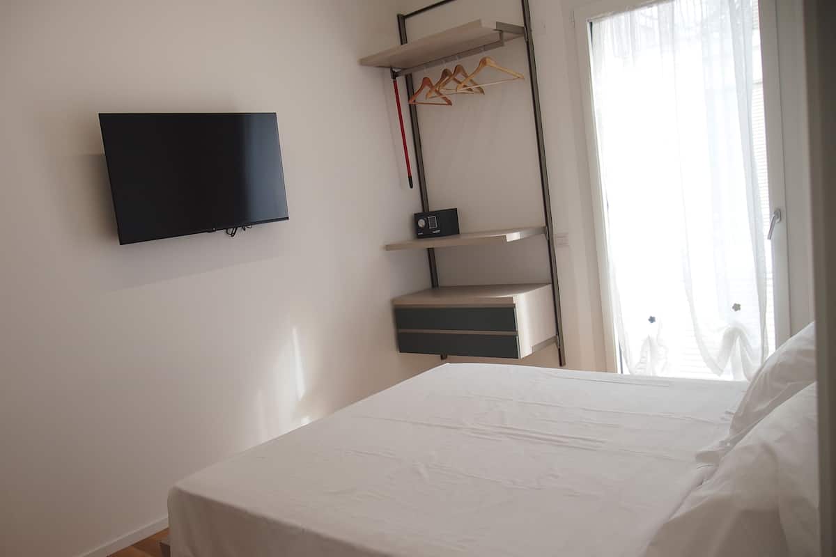 Quarto com cama, televisão fixada na parede, estante com roupas e janela com cortinas.