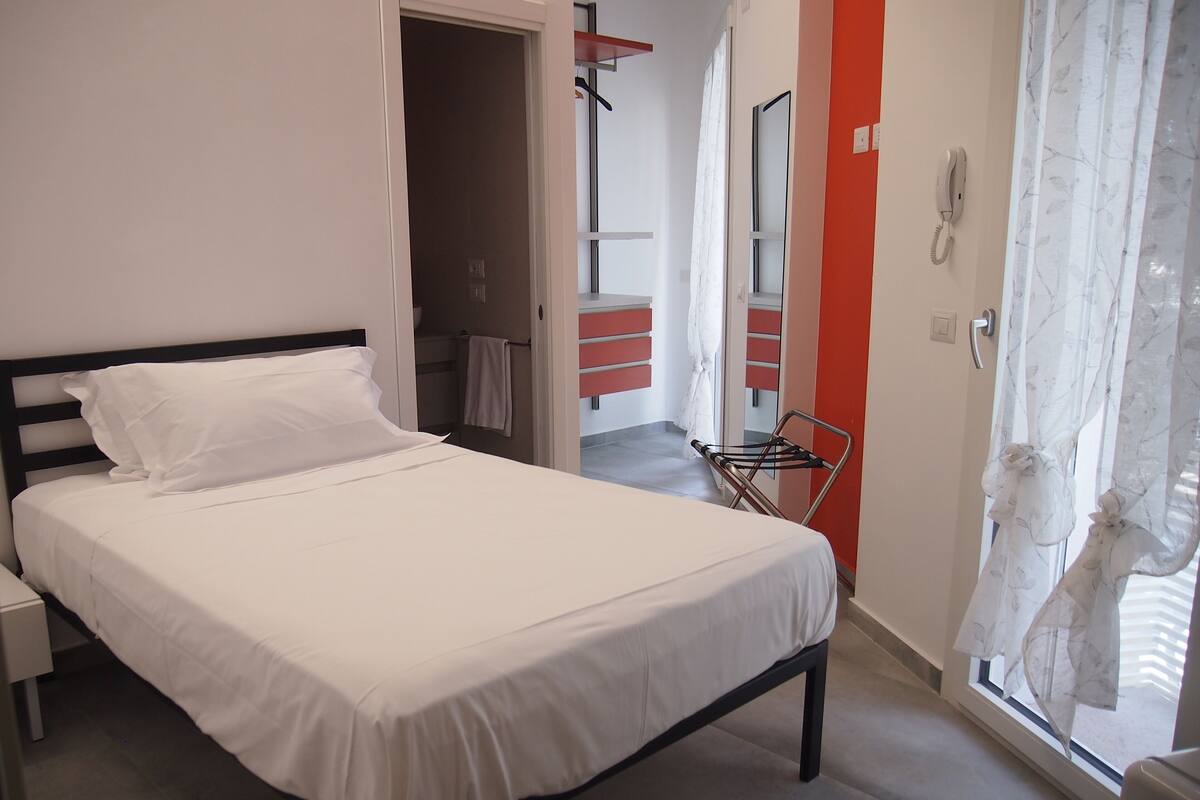 Cama bem arrumada em quarto com parede vermelha, telefone e janela com cortinas leves.