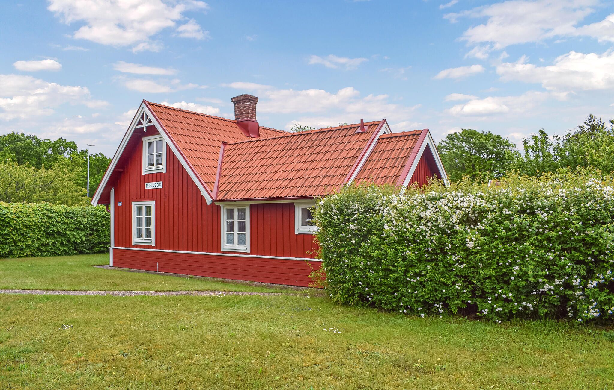 A red house with a sloped roof and a chimney, surrounded by greenery and a clear sky.