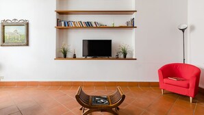 A living room with a TV on a shelf, a red chair, a wooden table, and a picture frame on the wall. - A 5 passi dalla Fontana di Trevi – Loft romano del XVII secolo! (Rome)