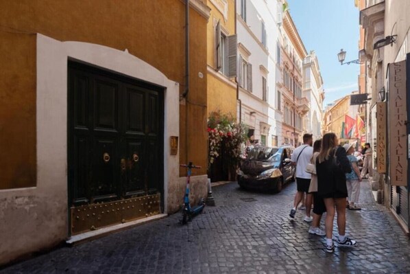 Una stretta strada acciottolata fiancheggiata da edifici, con un'auto nera parcheggiata a destra e un gruppo di persone che cammina.