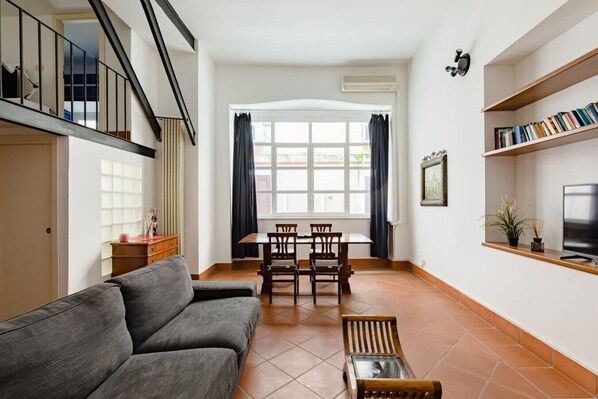 A living room with a sofa, a dining table with chairs, a TV on a shelf, and a window with curtains. - A 5 passi dalla Fontana di Trevi – Loft romano del XVII secolo! (Rome)