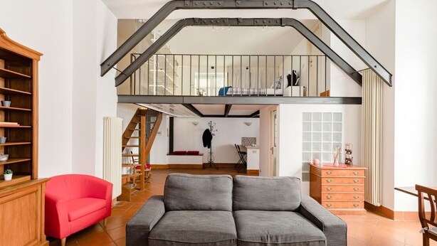 A spacious living room with a grey sofa, a red armchair, and a wooden cabinet. A staircase leads to an upper level. - A 5 passi dalla Fontana di Trevi – Loft romano del XVII secolo! (Rome)