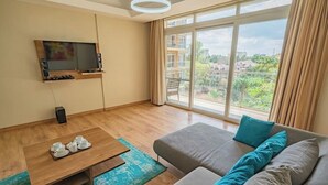 Een moderne woonkamer met een groot raam, een flatscreen televisie aan de muur, een grijze bank met blauwe kussens, en een houten salontafel met kopjes en schotels.