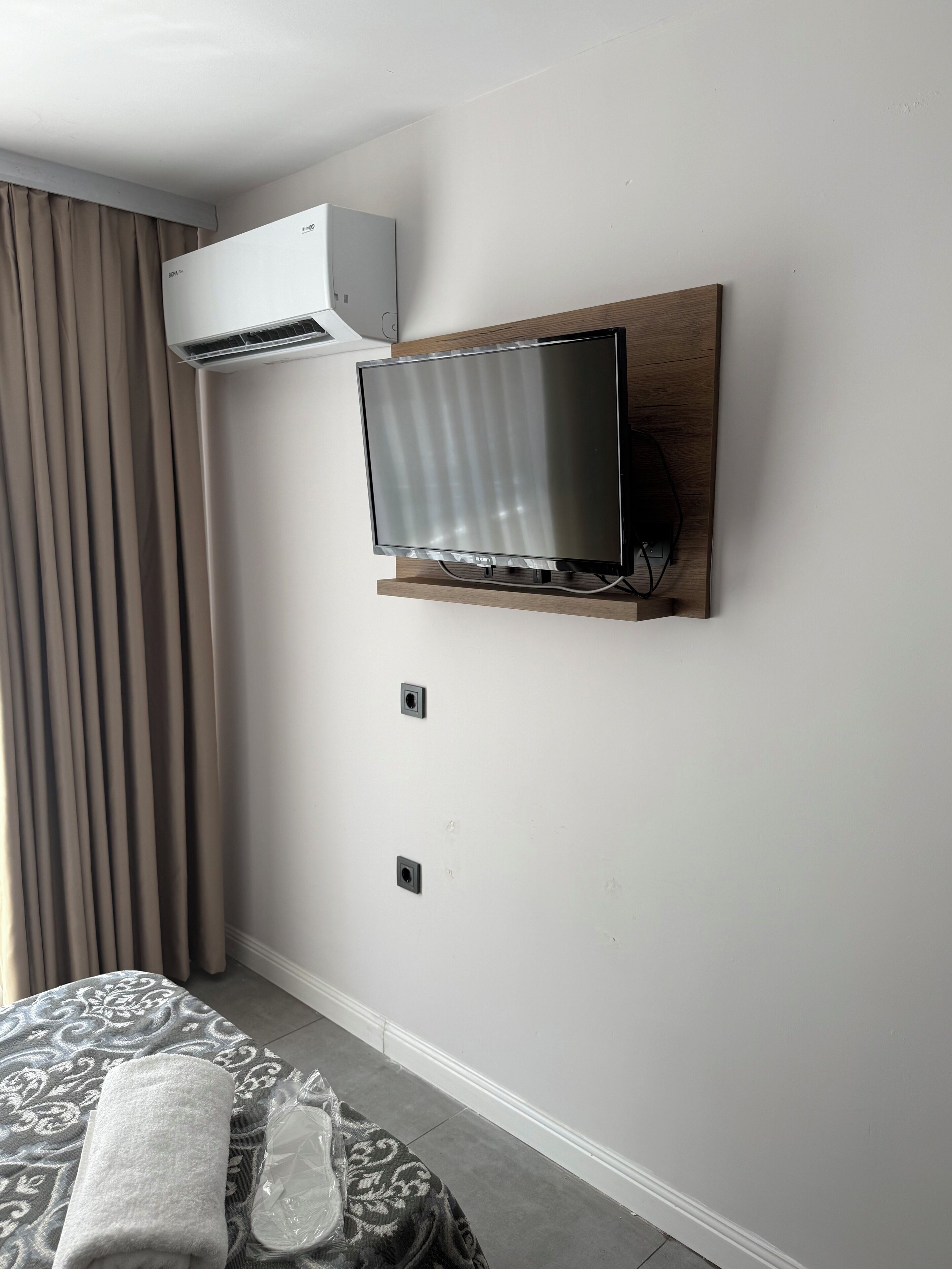 Une chambre d’hôtel équipée d’un téléviseur à écran plat fixé au mur, d’un appareil de climatisation, d’une étagère en bois et d’une couvre-lit à motifs.