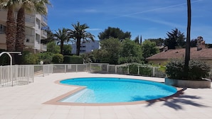 A swimming pool with a curved edge, surrounded by a white fence, palm trees, and a building with balconies in the background.