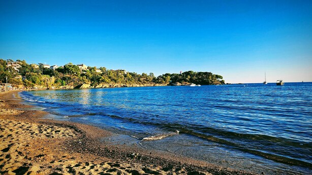 A beach with clear blue water, pebbly shore, and buildings in the background.