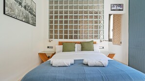 A neatly made bed with a blue bedspread, white pillows, and green accent pillows. There is a large window with a view of the city, and a framed picture on the wall.
