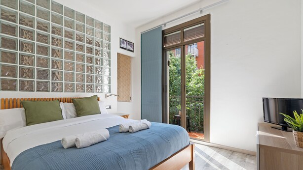 A modern hotel room with a large bed, a television, and a balcony with a view of greenery.