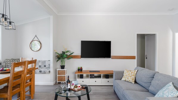 Un salon moderne avec un canapé gris, une table basse en verre, des chaises en bois, un téléviseur fixé au mur et un coin repas avec une nappe à motifs.