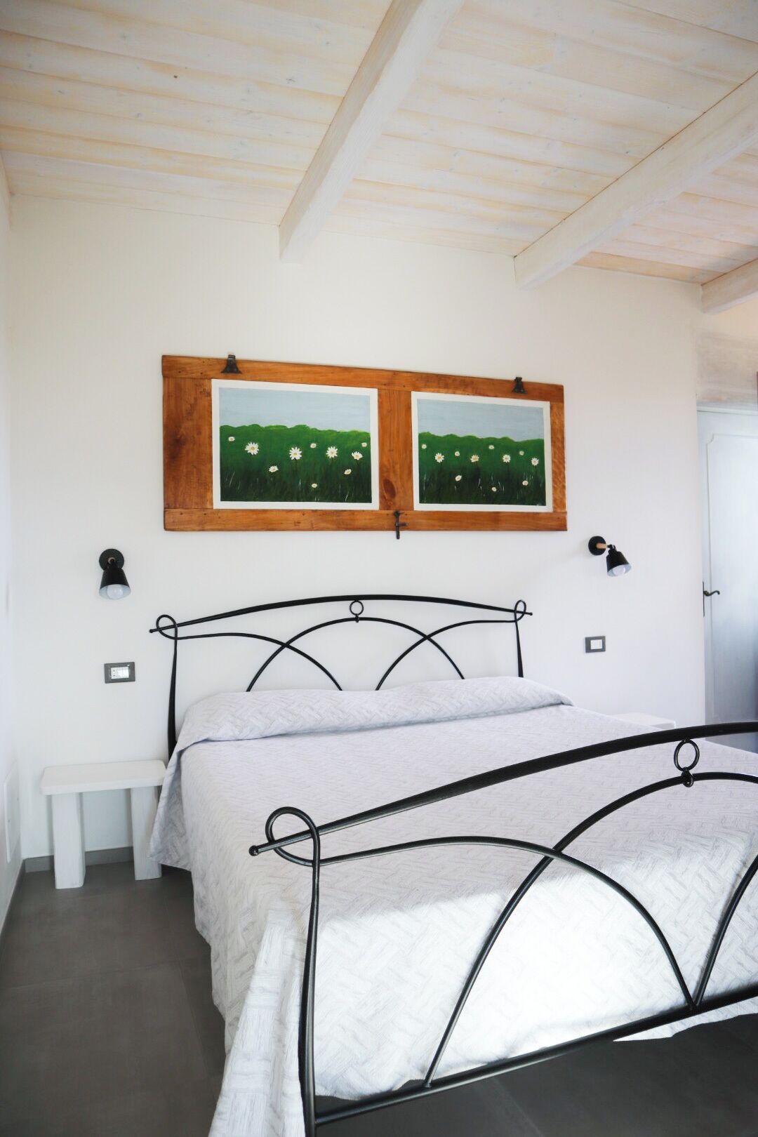 Un dormitorio con estructura de cama de metal, cabecera de madera, un banco y dos cuadros colgados en la pared.