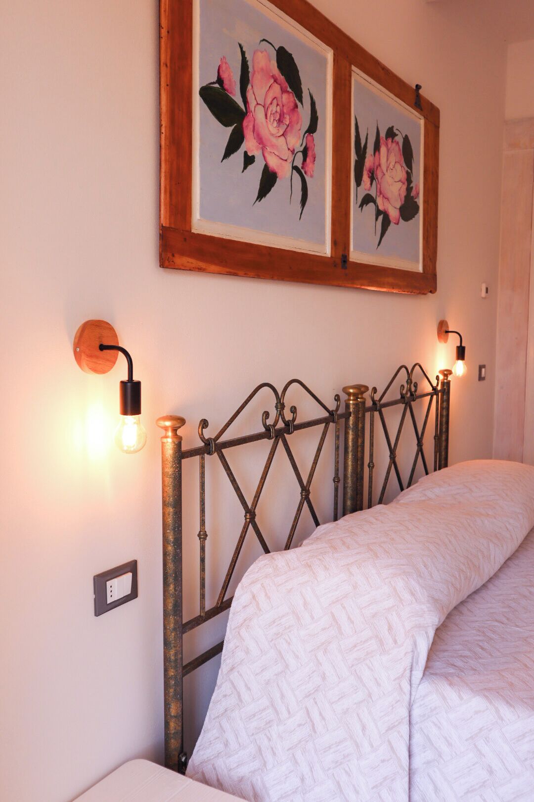 Una cama con cabecera de metal, dos lámparas de pared y dos cuadros con motivos florales.