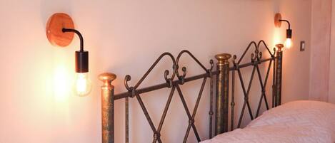 Una cama con cabecera de metal, dos lámparas de pared y dos cuadros con motivos florales.