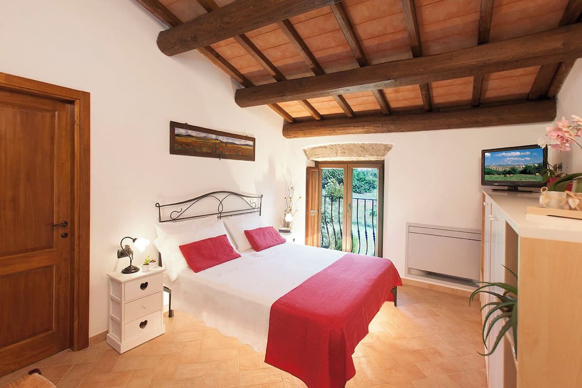 Quarto com cama, mesas de cabeceira, televisão e janela com vista para o verde.