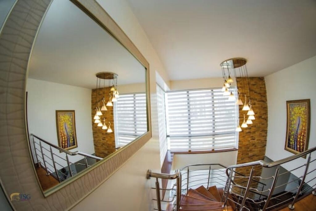 A staircase with metal railings and a wooden handrail, leading to a landing with a large mirror reflecting a chandelier and a painting.