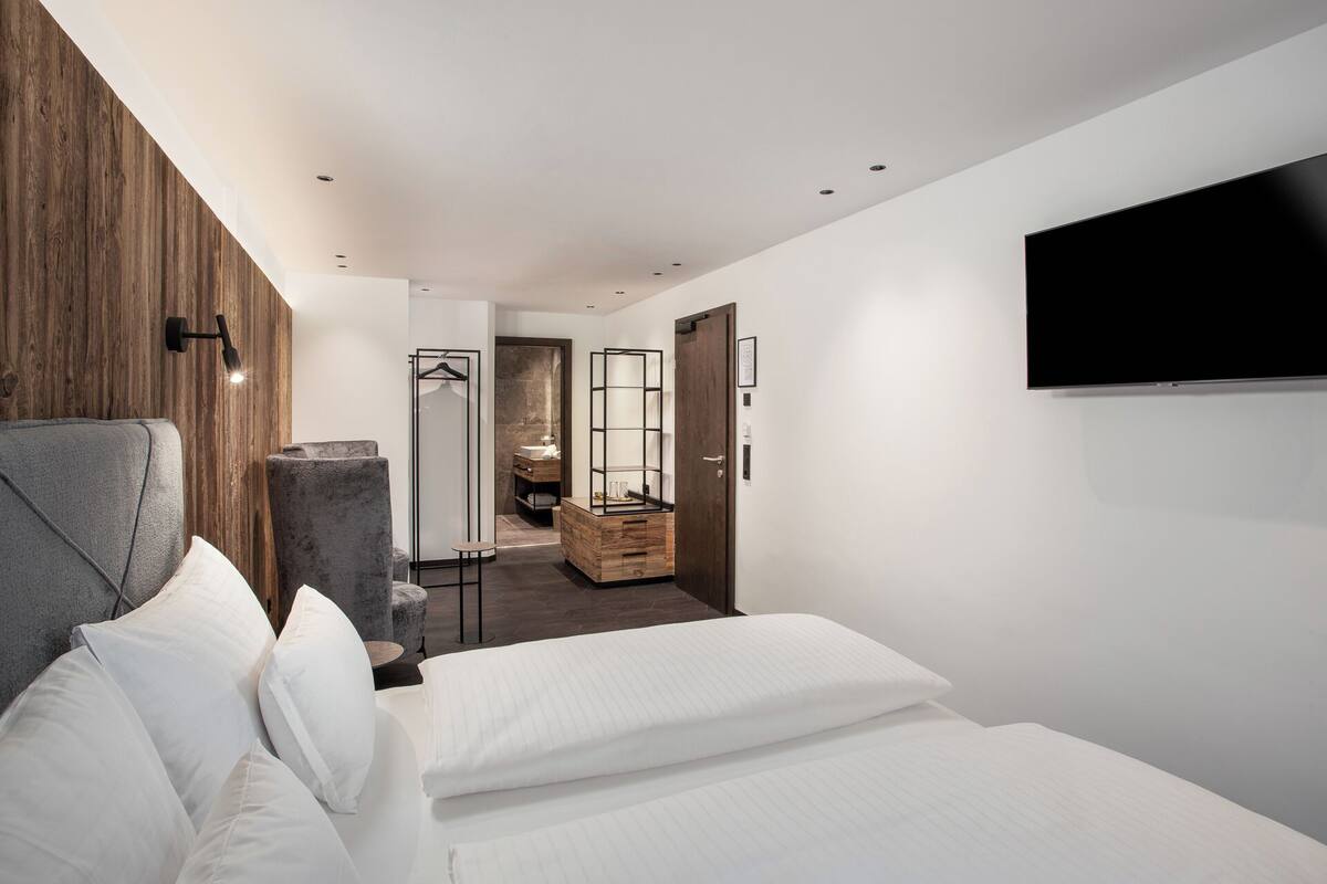 A modern hotel room with a bed, a TV, a wooden accent wall, a bathroom with a mirror and sink, and a hallway leading to another room.