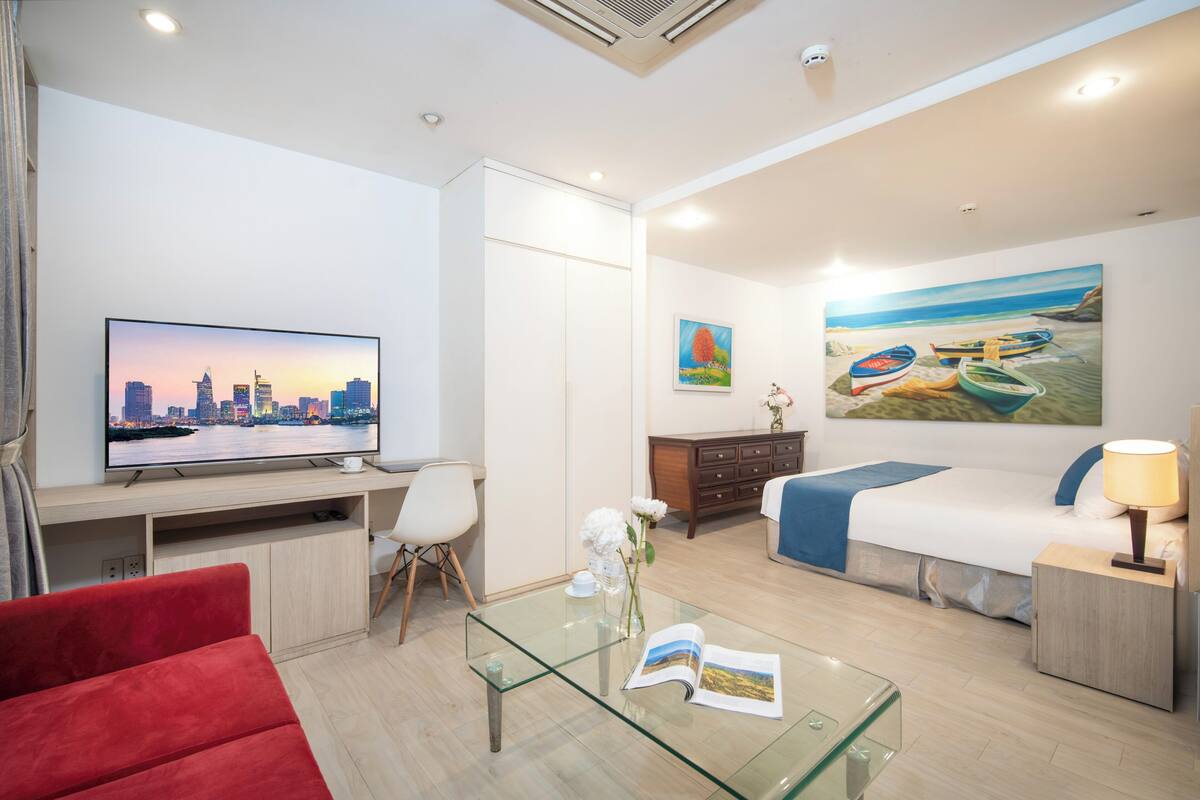 Un moderno cuarto de hotel con televisión mostrando un paisaje urbano, una cama con un cuadro de playa, una mesa de centro de vidrio y un sofá rojo.