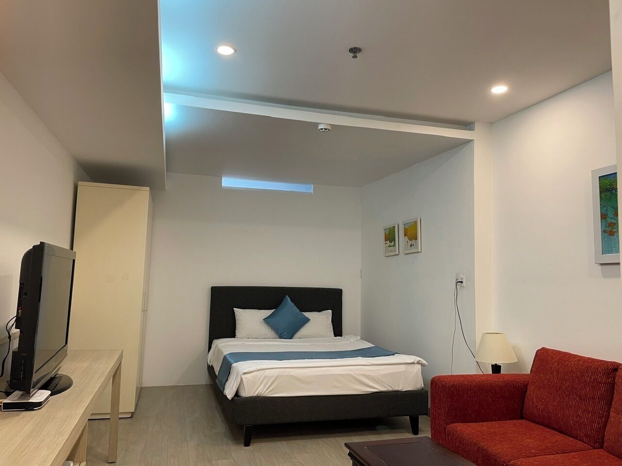 A hotel room with a bed, a TV, a desk, a red sofa, and a lamp.