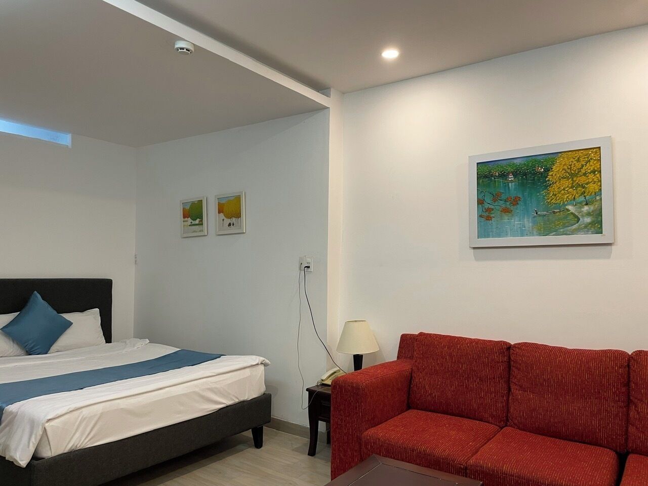 A bedroom with a bed, a red sofa, a nightstand with a lamp, and a painting on the wall.
