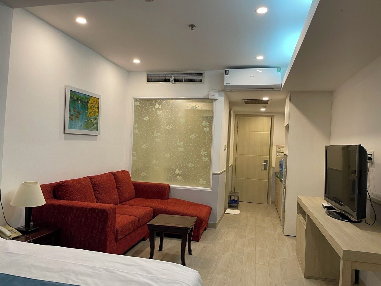 Une chambre d’hôtel avec un canapé rouge, une petite table, un téléviseur posé sur un meuble, un lit, une fenêtre avec des stores à motifs floraux et un couloir menant à une salle de bain.