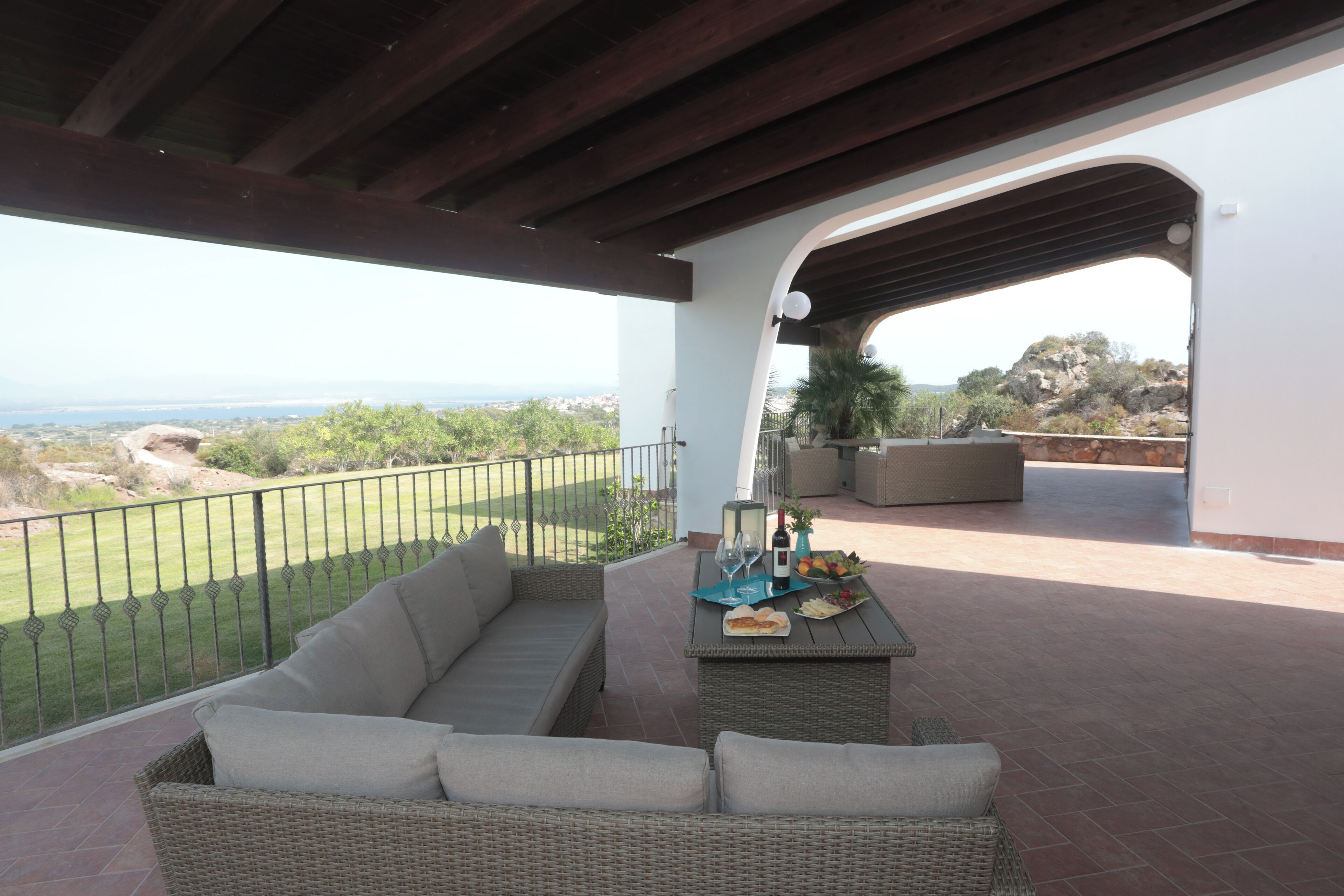 Um alpendre coberto com um sofá de vime, uma mesa com garrafa, copos e frutas, e vista para a paisagem.