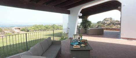 Um alpendre coberto com um sofá de vime, uma mesa com garrafa, copos e frutas, e vista para a paisagem.