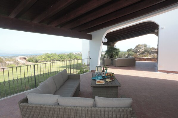 A covered patio with a wicker sofa, a table with a bottle, glasses, and fruit, and a view of the landscape. - rocce antiche resort (Sant'Antioco)