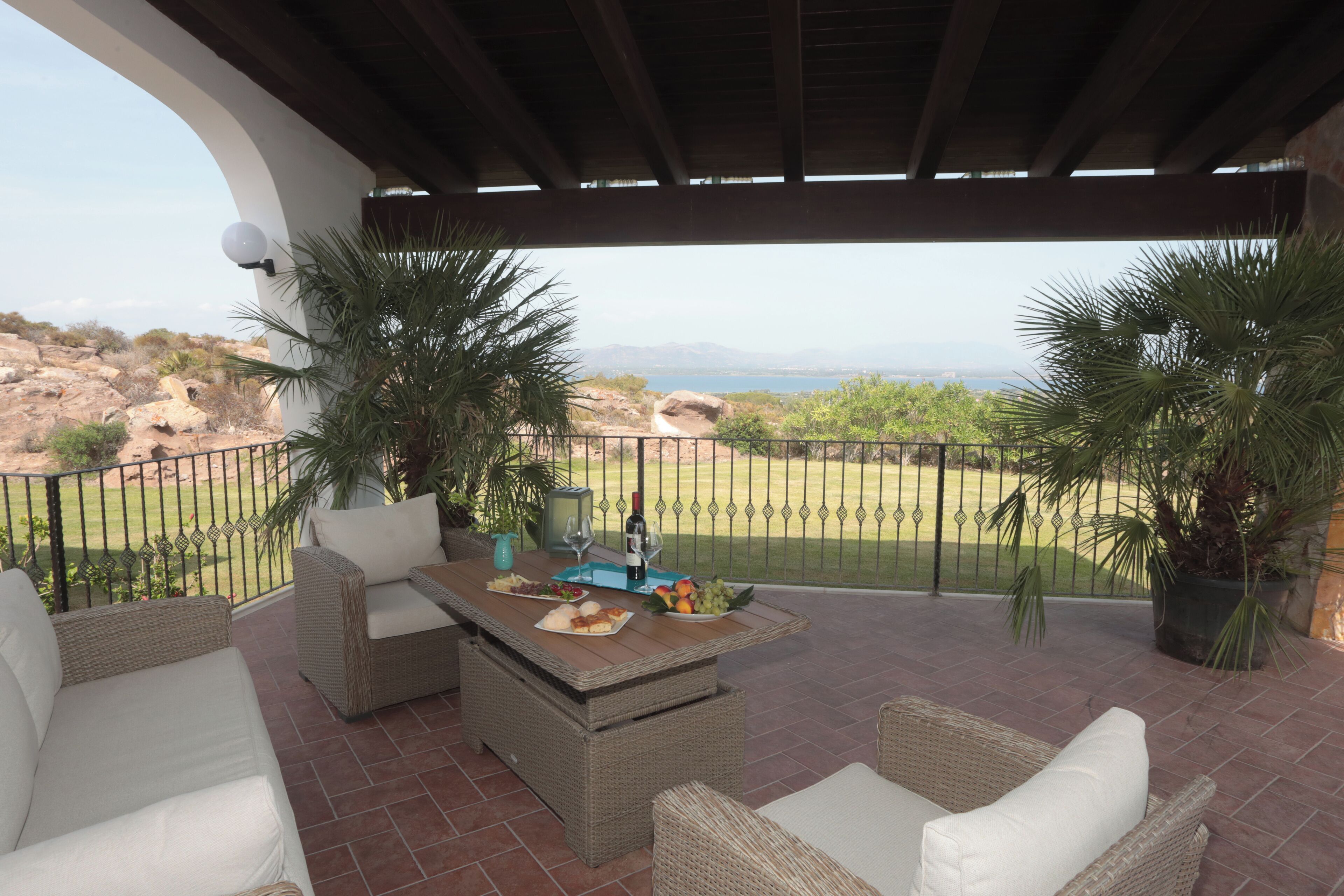 A covered patio with wicker furniture, a table set with food and drinks, and a view of the landscape.