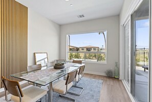 Un espace repas moderne avec une table en verre, des chaises rembourrées et une vue sur un quartier résidentiel grâce à de grandes fenêtres.