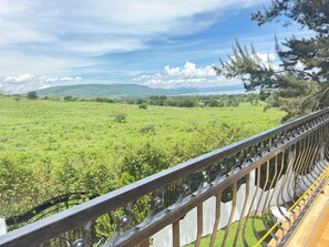 Una vista panorámica desde un balcón con baranda de metal, sobre un paisaje verde y exuberante con colinas lejanas y cielo despejado.