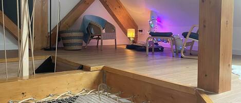 A loft bedroom with a sloped ceiling, wooden beams, a hammock, a small table, and a chair.
