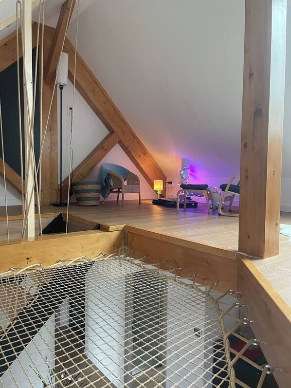 A loft bedroom with a sloped ceiling, wooden beams, a hammock, a small table, and a chair.