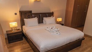 A neatly made bed with white linens and a set of towels on the bed. There are two bedside tables with lamps, a wooden headboard, and a wardrobe in the background.